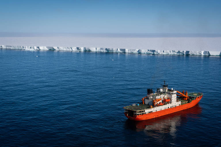 A imagem mostra um navio de pesquisa de cor laranja e branca navegando em águas azuis, com uma grande plataforma de gelo ao fundo. O céu está claro e sem nuvens, e a linha do horizonte é visível onde o gelo encontra o céu.