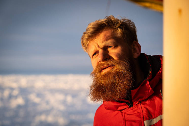 Um homem com barba longa e loira， usando um casaco vermelho， está olhando para fora de uma estrutura， com um fundo de gelo e céu claro. A luz do sol ilumina seu rosto， destacando sua expressão pensativa.