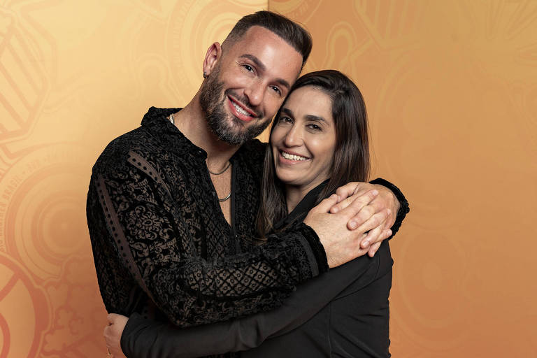 A imagem mostra um homem e uma mulher sorrindo e se abraçando. O homem está usando uma camisa preta com detalhes em veludo e tem barba e cabelo curto. A mulher está vestindo uma blusa preta e ambos estão em um fundo laranja com padrões sutis.