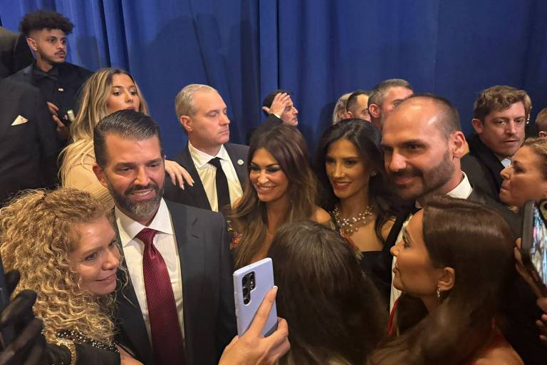 A imagem mostra um grupo de pessoas em um evento social， com um fundo azul. Algumas pessoas estão sorrindo e conversando， enquanto uma mão segura um celular， filmando ou tirando uma foto do grupo. A maioria das pessoas está vestida formalmente， com trajes como ternos e vestidos. O foco parece estar em um homem com cabelo escuro e barba， que está sorrindo.