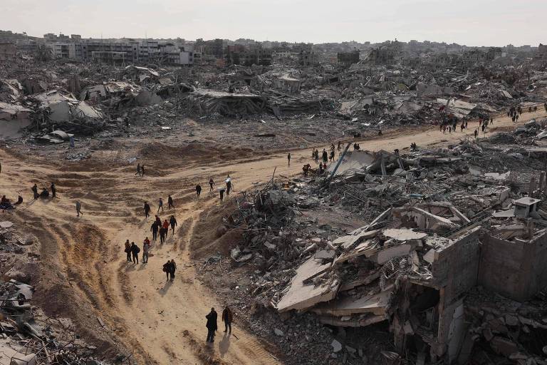 A imagem mostra uma área devastada， com muitos destroços e ruínas visíveis. No centro， há um caminho de terra batida que é percorrido por várias pessoas. O céu está nublado， e a paisagem é marcada por escombros e estruturas destruídas ao fundo.