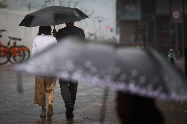 A imagem mostra um casal caminhando numa calçada molhada， ambos sob o mesmo guarda-chuvas. Em primeiro plano， desfocado， há outra pessoa passando com outro guarda-chuva que ocupa boa parte da imagem. A cena é em um ambiente urbano， com a presença de bicicletas ao fundo. A chuva é visível， com gotas caindo sobre os guarda-chuvas. A iluminação é suave， sugerindo um momento com sol encoberto.
