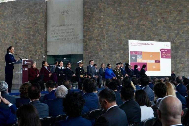 A imagem mostra um evento em um espaço interno com uma parede de pedra ao fundo. Uma mulher está em pé em um palco， falando ao microfone， enquanto uma audiência a observa. No palco， há várias pessoas sentadas， incluindo representantes de diferentes instituições. À direita， há uma tela com gráficos e informações visíveis. A plateia é composta por homens e mulheres， alguns usando ternos.