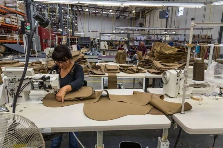 A imagem mostra uma mulher trabalhando em uma mesa de costura em uma fábrica. Ela está concentrada em cortar ou costurar peças de tecido， que parecem ser de um material marrom. Ao fundo， há várias máquinas de costura e materiais organizados， indicando um ambiente de produção.