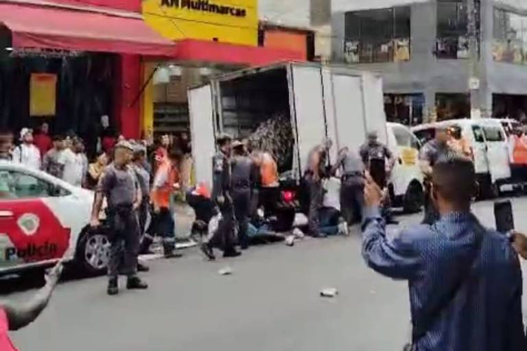 A imagem mostra uma cena em uma rua movimentada， onde várias pessoas estão reunidas em torno de um caminhão de entrega. Há uma presença significativa de policiais e dois homens no chão. Uma multidão observa a cena. O ambiente é urbano， com lojas e edifícios ao fundo 