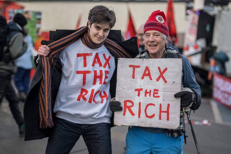 A  imagem mostra duas pessoas em um protesto. À esquerda， um jovem vestindo uma camiseta branca com a frase 039;TAX THE RICH039; em letras vermelhas. À direita， um homem mais velho usando um gorro vermelho e segurando uma placa com a mesma frase escrita em letras grandes. O fundo apresenta bandeiras e outras pessoas participando do protesto.