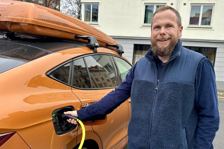 Um homem com barba e sorriso está ao lado de um carro elétrico laranja, segurando um cabo de carregamento que está conectado ao veículo. O carro possui um design moderno e está estacionado em uma rua com edifícios ao fundo.