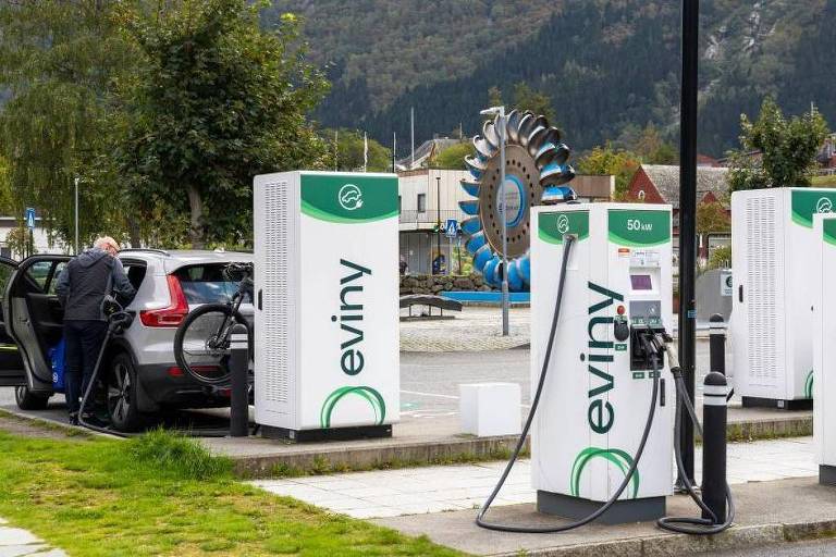 A imagem mostra uma estação de carregamento para veículos elétricos, com várias unidades de carregamento brancas e verdes da marca 'eviny'. No fundo, há uma roda-gigante e montanhas ao longe, indicando um ambiente urbano ou turístico. Alguns carros estão estacionados nas proximidades.