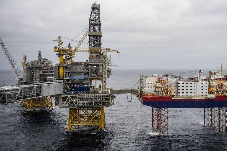 A imagem mostra duas plataformas de perfuração no mar, uma delas é uma plataforma fixa com estrutura amarela e a outra é uma plataforma flutuante com casco vermelho e branco. O céu está nublado e o mar parece agitado.