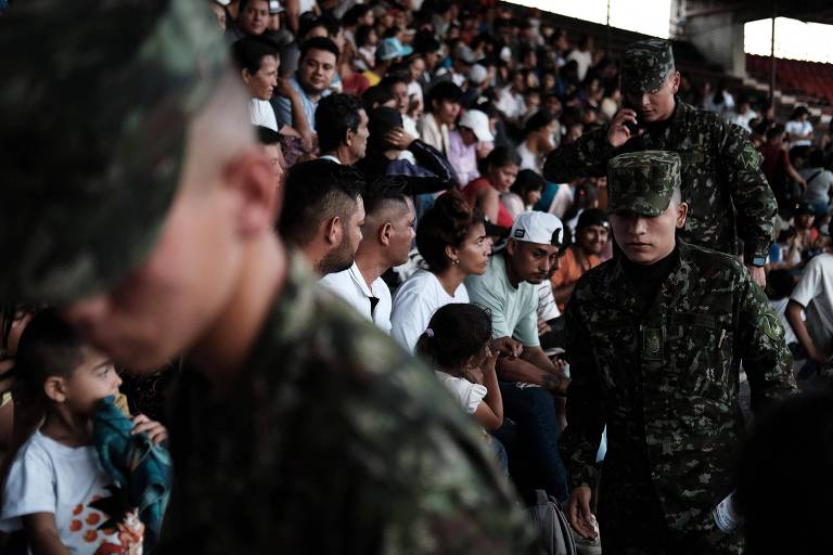 Imagem mostra militares trajados com roupas camufladas em primeiro plano e， ao fundo， uma multidão de pessoas sentadas na arquibancada de um estádio 