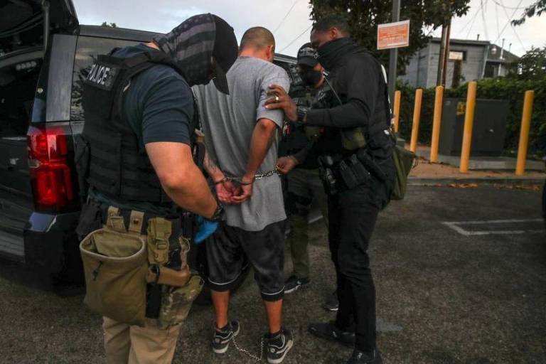 A imagem mostra um homem sendo algemado por policiais em uma área externa. O homem está de costas， vestindo uma camiseta cinza e shorts escuros. Dois policiais estão próximos a ele， um deles está usando um colete à prova de balas e o outro está com uma máscara. Um veículo policial está ao fundo， e há postes de luz e uma placa visível na área.