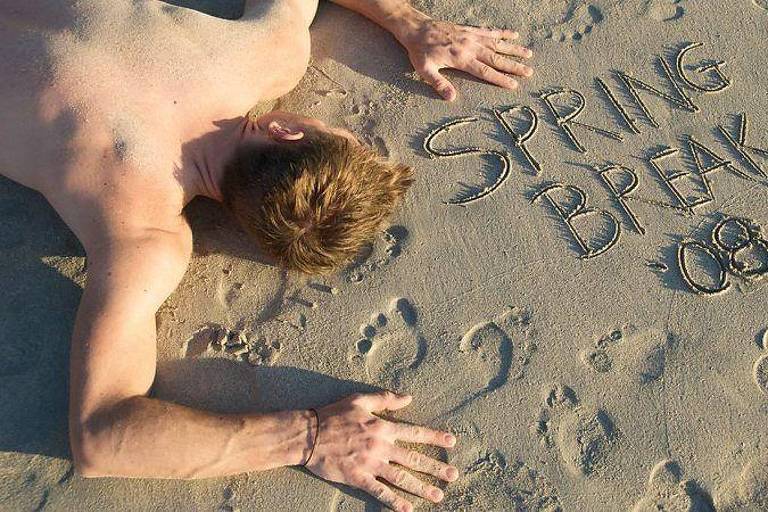 Homem sem camisa deitado de bruços na areia de uma praia parecendo estar desmaiado após ter bebido demais， ao lado dele está escrito na areia 039;Férias de Primavera 2008039; em inglês