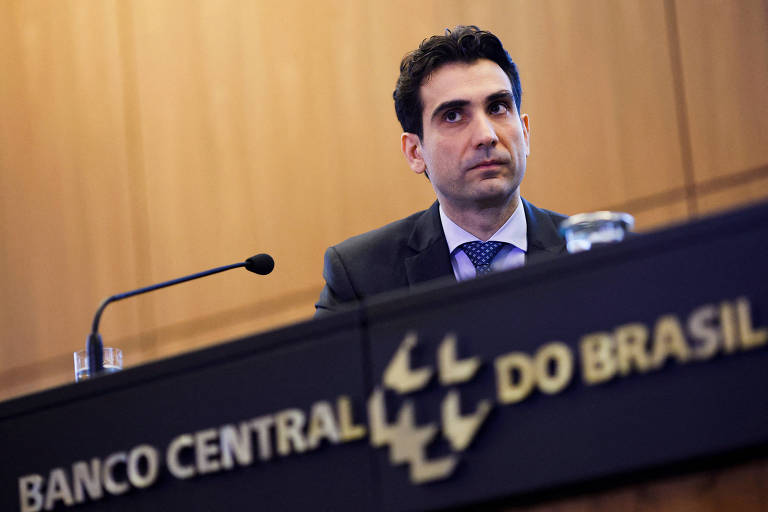 A imagem mostra um homem sentado em uma mesa durante uma reunião no Banco Central do Brasil. Ele está olhando para frente， com uma expressão séria. Ao fundo， há uma parede de madeira e um painel com o nome 039;BANCO CENTRAL DO BRASIL039;. Na mesa， há um copo de água e um microfone.
