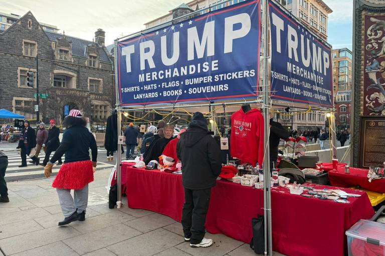 A imagem mostra uma barraca de mercadorias com o nome 039;TRUMP MERCHANDISE039; em destaque. A barraca oferece uma variedade de produtos， incluindo camisetas， chapéus， bandeiras， adesivos para para-choques， cordões， livros e souvenirs. Algumas pessoas estão ao redor da barraca， e uma delas está vestindo uma saia de palha vermelha. Ao fundo， há um edifício de estilo arquitetônico antigo e outras pessoas caminhando pela calçada.