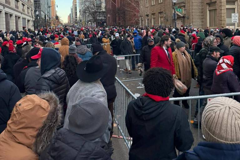 A imagem mostra uma grande multidão reunida em uma rua， com prédios altos ao fundo. As pessoas estão vestidas com roupas de inverno， algumas usando gorros e cachecóis. A maioria das pessoas parece estar usando roupas escuras， com algumas destacando-se em vermelho. Há barreiras de segurança visíveis na frente da multidão， e o céu está claro.