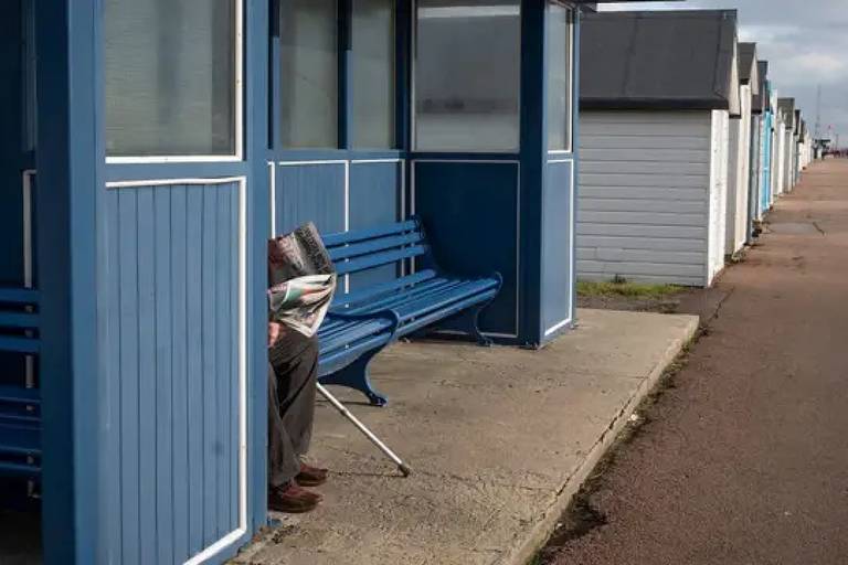 A imagem mostra uma fileira de cabines de praia de cor azul e branca. À esquerda, uma pessoa está sentada em um banco, parcialmente escondida atrás de uma cabine, lendo um jornal. O chão é de concreto e há uma calçada ao lado das cabines.