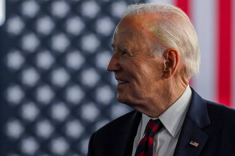 A imagem mostra um homem de perfil， com cabelo grisalho e um sorriso. Ele está usando um terno escuro e uma gravata listrada em vermelho e preto. Ao fundo， há uma bandeira dos Estados Unidos com listras vermelhas e brancas e um padrão de estrelas brancas sobre um fundo azul.