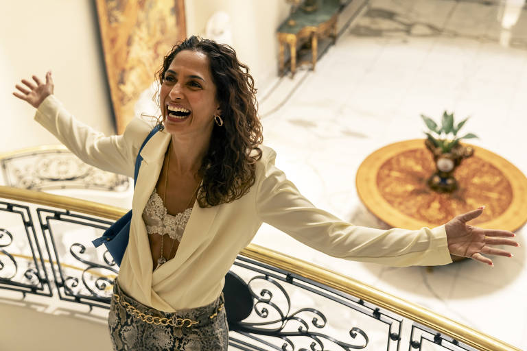 Uma mulher com cabelo cacheado e solto, vestindo um blazer branco e uma blusa de renda, está em uma escada com os braços abertos, sorrindo. Ao fundo, há um ambiente decorado com um tapete e uma mesa redonda. O piso é de mármore e há uma planta em cima da mesa.