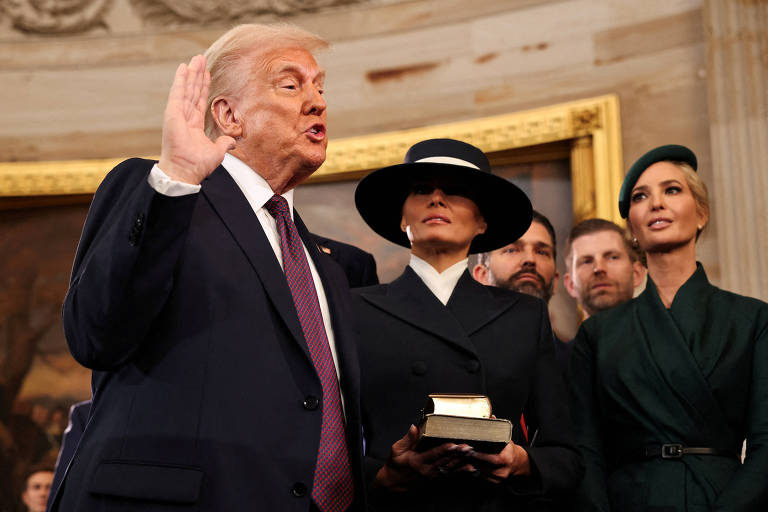 A imagem mostra Donald Trump em um terno escuro levantando a mão direita, fazendo um juramento. Ao seu lado, uma mulher com um chapéu preto e um vestido branco segura um livro. Atrás deles, há outras pessoas, incluindo um homem com cabelo loiro e uma mulher com cabelo claro, vestindo um vestido verde. O fundo apresenta uma decoração clássica com uma moldura dourada.
