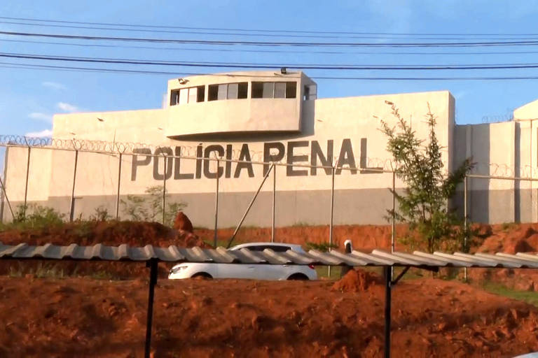 A imagem mostra um grande edifício com a inscrição 039;POLICIA PENAL039; em letras grandes e visíveis. O prédio é cercado por uma cerca e há um pequeno edifício de observação no topo. O céu está claro e há algumas árvores ao redor. No primeiro plano， há uma área de terra e um carro estacionado.
