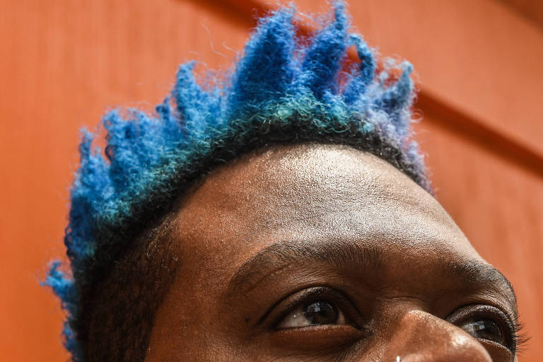 A imagem mostra a parte superior da cabeça de uma pessoa com cabelo estilizado em um tom azul vibrante， com algumas mechas em azul claro. O fundo é uma parede laranja， que contrasta com a cor do cabelo.