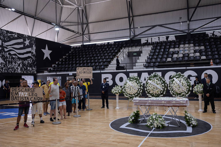 A imagem mostra um ginásio esportivo onde está sendo realizada uma homenagem. No centro， há um caixão coberto com flores brancas， cercado por arranjos florais. À frente do caixão， um grupo de pessoas está em fila， segurando cartazes com mensagens de despedida. O ambiente é decorado com as cores do time， e as arquibancadas estão vazias. Ao fundo， há uma bandeira e o nome do ginásio visível na parede.
