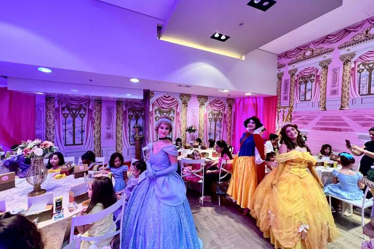 A imagem mostra uma festa temática de princesas em um ambiente decorado com paredes que imitam um castelo. Três personagens femininas vestidas como princesas estão em destaque: uma com um vestido azul claro (Cinderela), outra com um vestido amarelo (Bela) e uma terceira com um vestido vermelho e amarelo (Branca de Neve). Várias crianças estão sentadas em mesas decoradas, algumas comendo e outras interagindo. O ambiente é iluminado com luzes suaves e há flores em um vaso central na mesa.