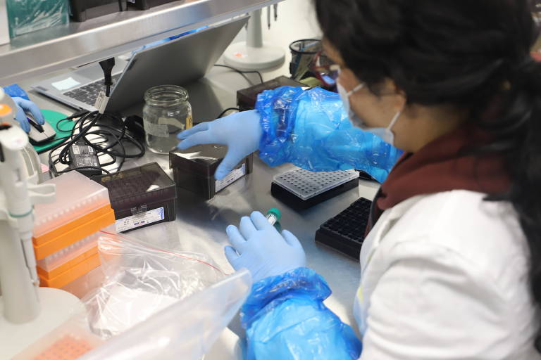 Uma cientista está trabalhando em um laboratório. Ela usa luvas azuis e uma jaqueta branca. A cientista está segurando um copo preto e parece estar manipulando substâncias. Ao fundo， há equipamentos de laboratório， incluindo um computador e frascos.