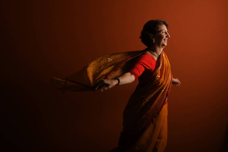 A imagem mostra uma mulher usando um sari laranja， com uma blusa vermelha， girando com os braços abertos. O fundo é de uma cor laranja sólida， criando um contraste vibrante. A mulher tem um sorriso no rosto e parece estar se divertindo.