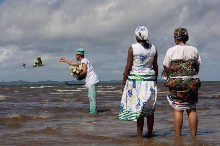 A imagem mostra três pessoas em uma praia. Duas mulheres estão de costas, vestindo roupas brancas e coloridas, enquanto um homem, de frente, está lançando flores ao mar. O céu está parcialmente nublado e as ondas do mar estão visíveis. Ao fundo, há uma pequena embarcação.