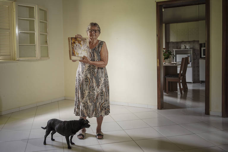 Uma mulher idosa está em pé em um ambiente interno， segurando uma revista ou livro. Ela usa um vestido longo com estampa de animal e óculos. Ao seu lado， há um cachorro preto. O ambiente é iluminado， com paredes claras e um piso de cerâmica. Ao fundo， pode-se ver uma cozinha e uma mesa.