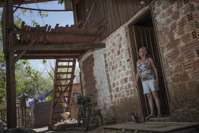 Uma mulher está de pé na entrada de uma casa de tijolos expostos. Ela usa uma blusa de alças e shorts brancos. Ao fundo， há uma escada de madeira que leva a um andar superior. O ambiente é rural， com vegetação ao redor e uma bicicleta encostada na parede.