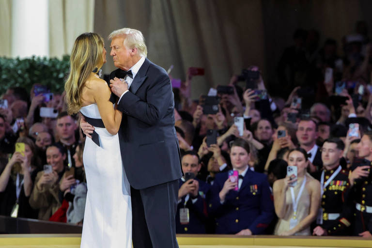 Donald Trump， homem branco com pele de tom alaranjado e cabelos grisalhos， usa um terno preto. Está em pé ao centro de um palco abraçado com sua mulher， Melania Trump， a quem segura pela cintura. Melania é uma mulher， branca， cabelos loiros. Ela aparece de costas para a câmera que registrou a fotografia. Usa um vestido branco com decote nas costas. Ao fundo há uma  plateia de pessoas que observam atentamente o casal 