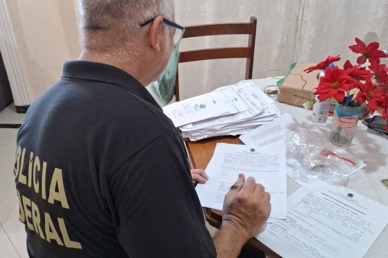 A imagem mostra um homem de costas， usando uma camisa preta com a inscrição 039;POLÍCIA FEDERAL039;. Ele está sentado à mesa， escrevendo em um documento. Ao redor dele， há várias folhas de papel empilhadas e um vaso com flores vermelhas. O ambiente parece ser uma sala bem iluminada.