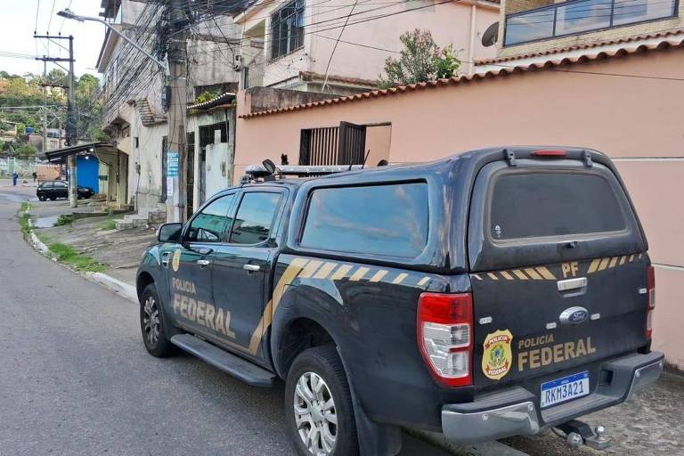 A imagem mostra um veículo da polícia estacionado em uma rua residencial. O carro é uma caminhonete com a pintura característica da polícia， incluindo emblemas e faixas. Ao fundo， há casas e postes de eletricidade， com uma rua pavimentada que se curva à esquerda.