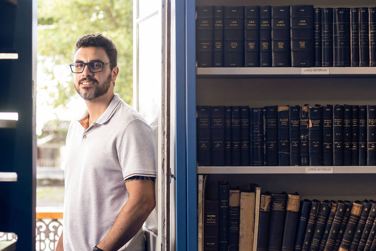 Um homem está posicionado ao lado de uma estante de livros em uma biblioteca. Ele usa uma camiseta clara e óculos， e está olhando para a câmera. Ao fundo， há uma estante com livros encadernados em capas escuras. A luz natural entra pela porta ao lado， iluminando o ambiente.