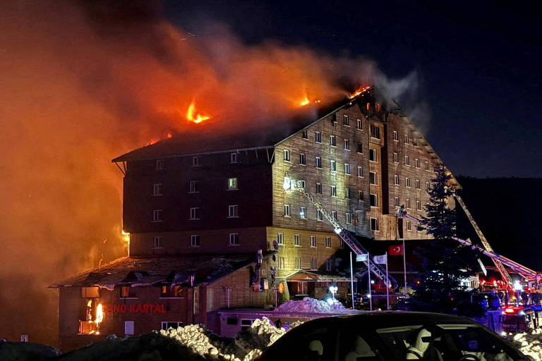 A imagem mostra um hotel em chamas durante a noite. O fogo está visível no telhado e nas janelas superiores， com fumaça densa subindo. No primeiro plano， há um veículo estacionado e， ao fundo， várias viaturas de bombeiros estão posicionadas， com mangueiras direcionadas para o incêndio. A cena é iluminada pela luz do fogo e das luzes de emergência.