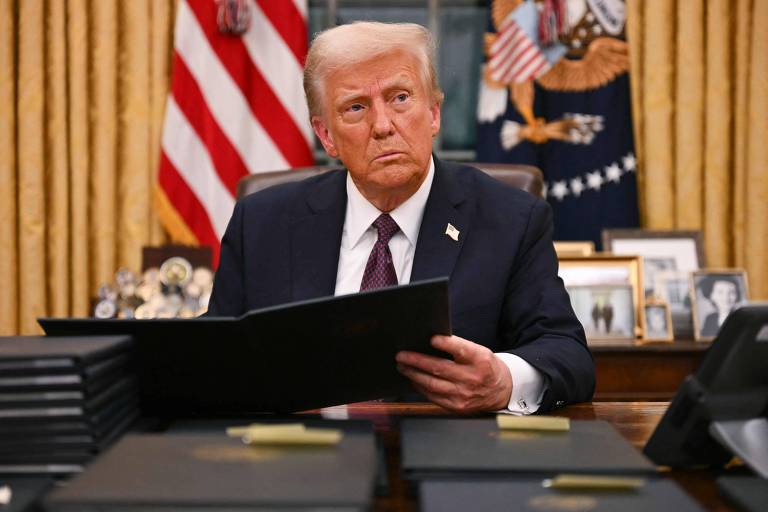 Um homem sentado em uma mesa no Salão Oval， com bandeiras dos Estados Unidos ao fundo. Ele segura um documento e parece pensativo. A mesa está cheia de pastas e há fotos emolduradas ao fundo.
