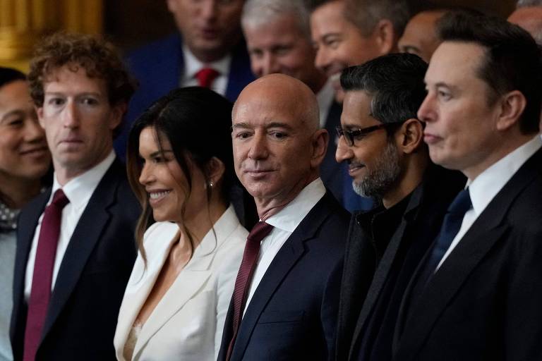 A imagem mostra um grupo de pessoas posando para a foto em um evento. No primeiro plano, há cinco indivíduos em destaque, com expressões sérias. Eles estão vestidos formalmente, com um homem à esquerda usando uma camisa e um blazer escuro, uma mulher com uma roupa branca, e outros homens em trajes escuros. Ao fundo, há mais pessoas visíveis, mas não estão em foco.
