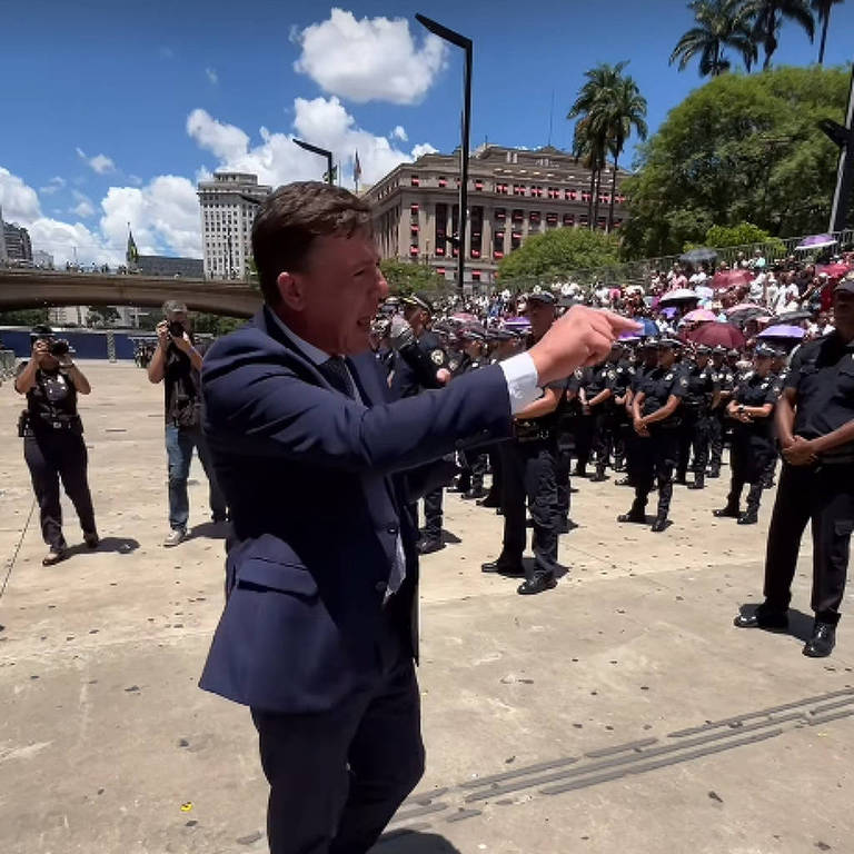 A imagem mostra um homem em um terno escuro， gesticulando enquanto fala para um grupo de pessoas. Ao fundo， há vários agentes de segurança em uniformes， alinhados em uma área externa com árvores e um edifício visível. O céu está claro e ensolarado.