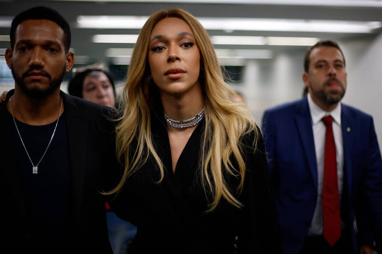 A imagem mostra um grupo de três pessoas em um ambiente interno. À frente， uma mulher com cabelo longo e liso， vestindo uma roupa escura e um colar， parece estar liderando o grupo. À esquerda， um homem com cabelo curto e barba， usando uma camiseta preta e um colar， observa atentamente. À direita， um homem em um terno azul com uma gravata vermelha também está presente. Ao fundo， outras pessoas são visíveis， mas não estão claramente focadas.
