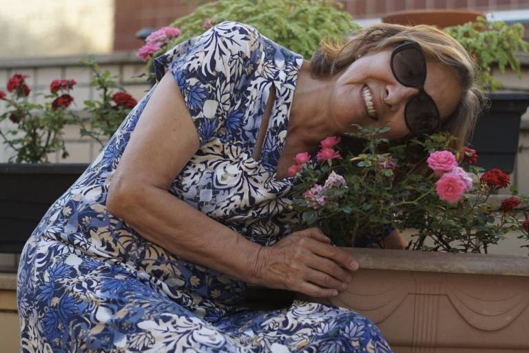 Uma mulher está agachada ao lado de um vaso de flores， com um vestido estampado em azul e branco. Ela parece estar acariciando as flores， com um sorriso no rosto. O fundo mostra um ambiente externo com mais plantas e flores.