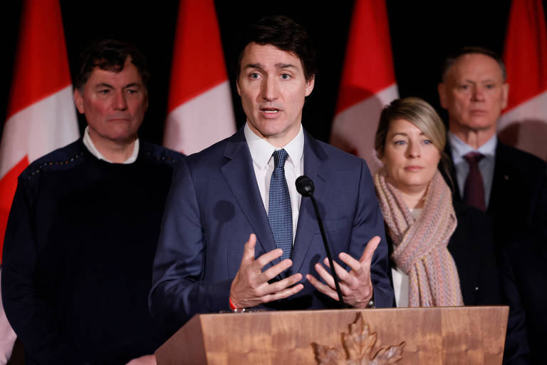 Imagem mostra Justin Trudeau， um homem em um terno escuro que fala em um microfone， gesticulando com as mãos. Atrás dele， há três pessoas: um homem à esquerda com um suéter escuro， uma mulher à direita com cabelo curto e um homem à direita com cabelo grisalho. Ao fundo， há bandeiras do Canadá.