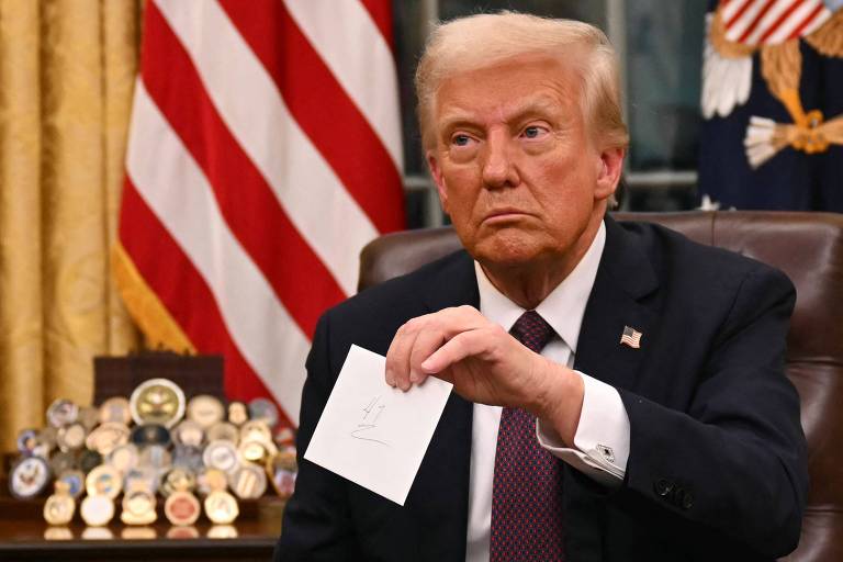 Um homem sentado em uma cadeira， segurando um papel em uma mão. Ele tem cabelo loiro e está vestido com um terno escuro e uma gravata. Ao fundo， há uma bandeira dos Estados Unidos e uma mesa com vários objetos， incluindo medalhas. O ambiente parece ser um escritório.