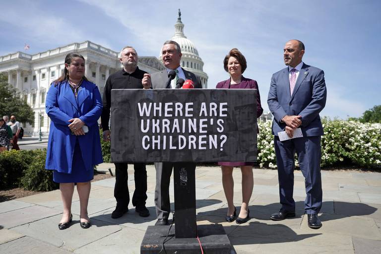 Um grupo de cinco pessoas está em pé em frente a um cartaz que diz 039;WHERE ARE UKRAINE039;S CHILDREN?039; (ONDE ESTÃO AS CRIANÇAS DA UCRÂNIA?). Ao fundo， pode-se ver o edifício do Capitólio dos Estados Unidos sob um céu claro. As pessoas estão vestidas de maneira formal， com algumas usando roupas em tons de azul e cinza.
