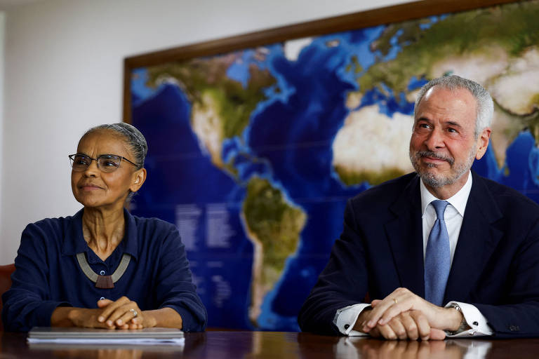 Marina e André com expressão sorridente sentados lado a lado em uma mesa; ao fundo há um mapa-múndi na parede