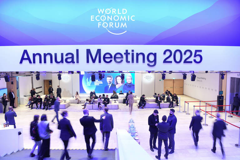 A imagem mostra um espaço de evento com o título 039;Annual Meeting 2025039; em destaque. O ambiente é amplo， com várias pessoas circulando e conversando. Ao fundo， há um painel com imagens de pessoas， e o logotipo do 039;World Economic Forum039; está visível no topo. O local parece ser bem iluminado e organizado， com mesas e cadeiras dispostas para os participantes.