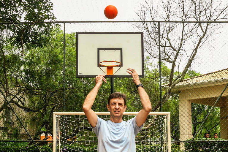 Um homem está em uma quadra de basquete， prestes a arremessar uma bola laranja em direção à cesta. A cesta está posicionada em uma estrutura metálica， com uma rede visível. Ao fundo， há árvores e um edifício. O homem usa uma camiseta clara e parece concentrado no arremesso.