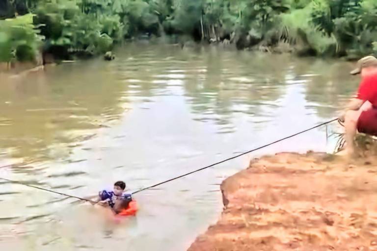 A imagem mostra uma criança em um pequeno barco flutuando em um rio. A criança está segurando uma corda que a conecta a um adulto sentado na margem do rio. O ambiente é natural， com vegetação ao redor e a água do rio calma.