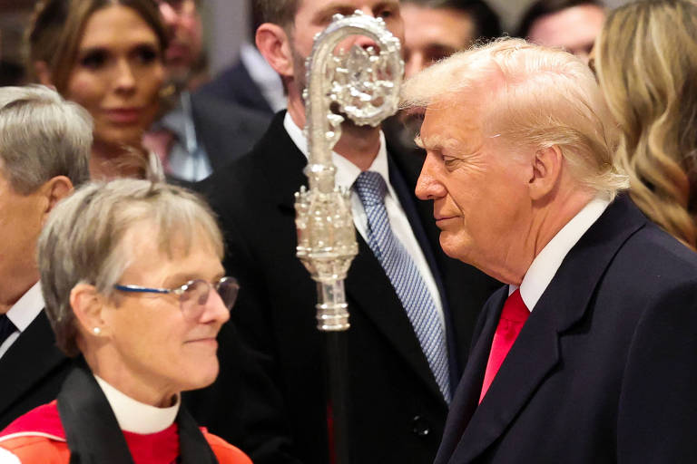 A imagem mostra um momento de interação entre duas pessoas em um evento cerimonial. À esquerda， uma mulher com óculos e vestindo uma vestimenta clerical está olhando para um homem à direita， que está usando um terno escuro e uma gravata vermelha. Ao fundo， há várias pessoas observando a cena. Um objeto prateado， possivelmente um cetro， está em destaque entre os dois